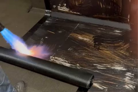 A lit gas torch heating the edge of a roll of membrane on a roof.
